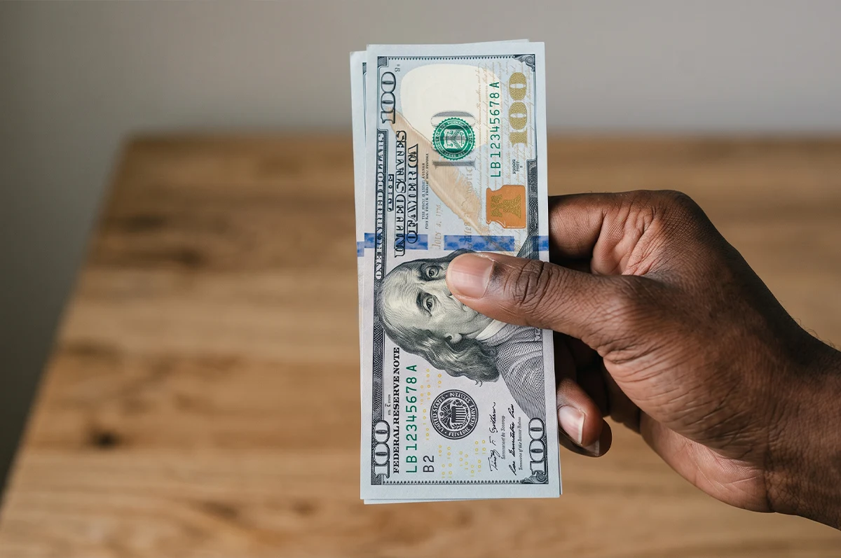 A person with dark skin holds a stack of crisp one-hundred-dollar bills vertically against a blurred, light-colored wooden table background surface