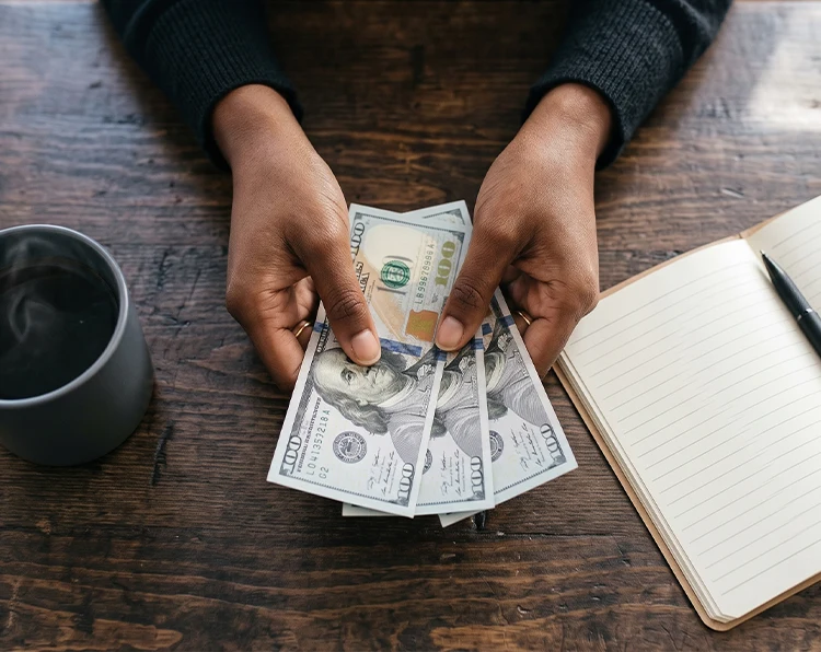 A woman's hands holding a few dollar bills on a table beside a coffee cup and a notebook